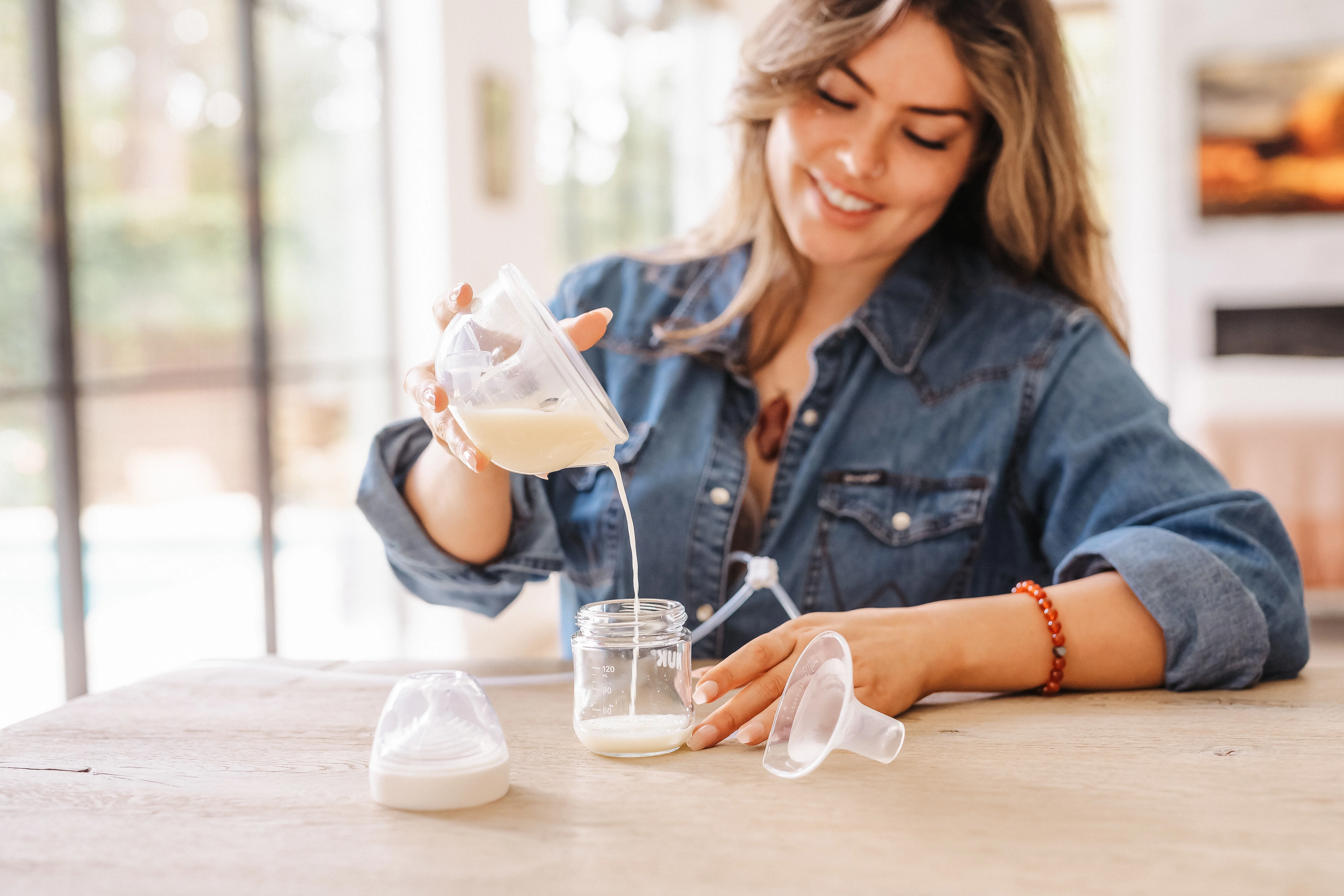 Mother pouring breast milk from a Legendairy Milk silicone collection cup into a glass storage jar at a kitchen table